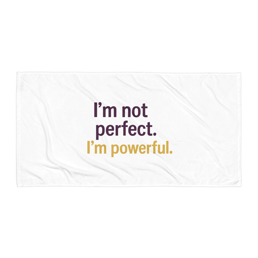 White beach towel with bold statement: “I’m not perfect. I’m powerful.” in purple and gold text on flat surface.