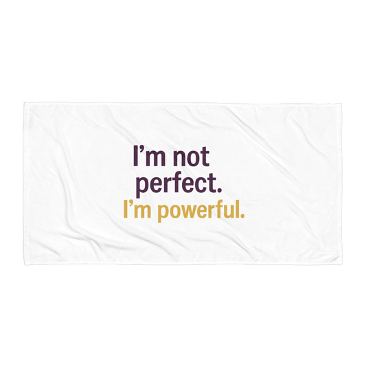White beach towel with bold statement: “I’m not perfect. I’m powerful.” in purple and gold text on flat surface.
