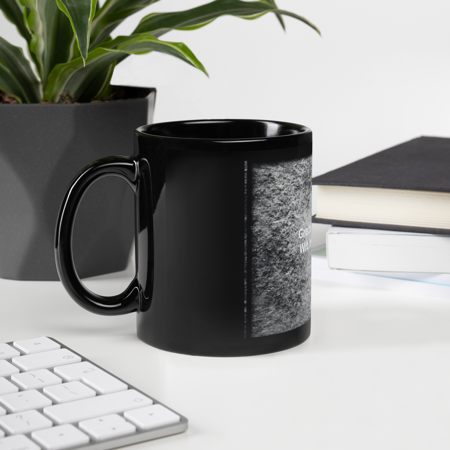 Black glossy mug on desk beside books and keyboard, featuring the quote. Grace in every sip. Wit in every drop design.
