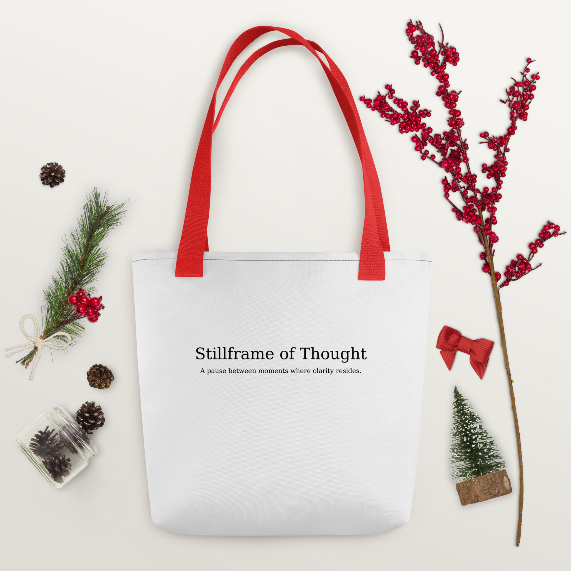 White tote bag with red handles and printed quote “Stillframe of Thought,” surrounded by pinecones and festive greenery.